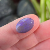 Blue oval gemstone held between fingers with a blurred natural background