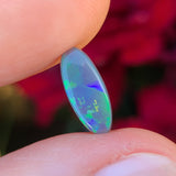 A small, elongated oval-shaped dark opal gemstone held between fingers, with a play of green color and a golden pattern reminiscent of music notes.